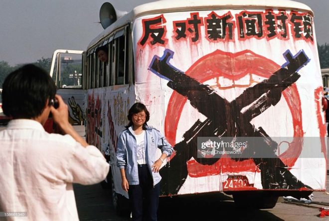 BEIJING, CHINA - 1989/06/01: A man takes a photograph of a woman standing next to a bus painted with protest slogans parked in Tiananmen Square. Pro-democracy demonstrators and protestors filled the square for weeks prior to the final nighttime Communist Government's bloody crackdown. Many camped out and slept in the square to keep the democracy protest movement going, while others, not directly involved in the protests, simply came by to see what was happening.. (Photo by Peter Charlesworth/LightRocket via Getty Images)